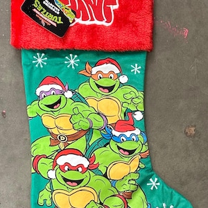 May include: Green Christmas stocking featuring the Teenage Mutant Ninja Turtles wearing Santa hats. The stocking has a red cuff and a green loop for hanging. The Turtles are depicted in a cartoon style with snowflakes.