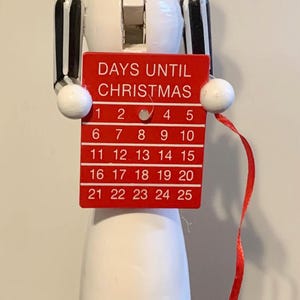 May include: A wooden snowman Christmas countdown calendar. The snowman has a green top hat, a painted face, and holds a red sign that reads "DAYS UNTIL CHRISTMAS" with numbered days. The base is red with glitter.
