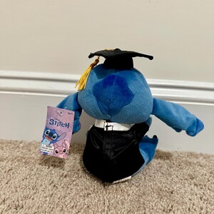 May include: A blue Stitch plush toy dressed in a black graduation gown and mortarboard with a gold tassel. The toy holds a tag with the Stitch logo and images of Stitch and Angel.