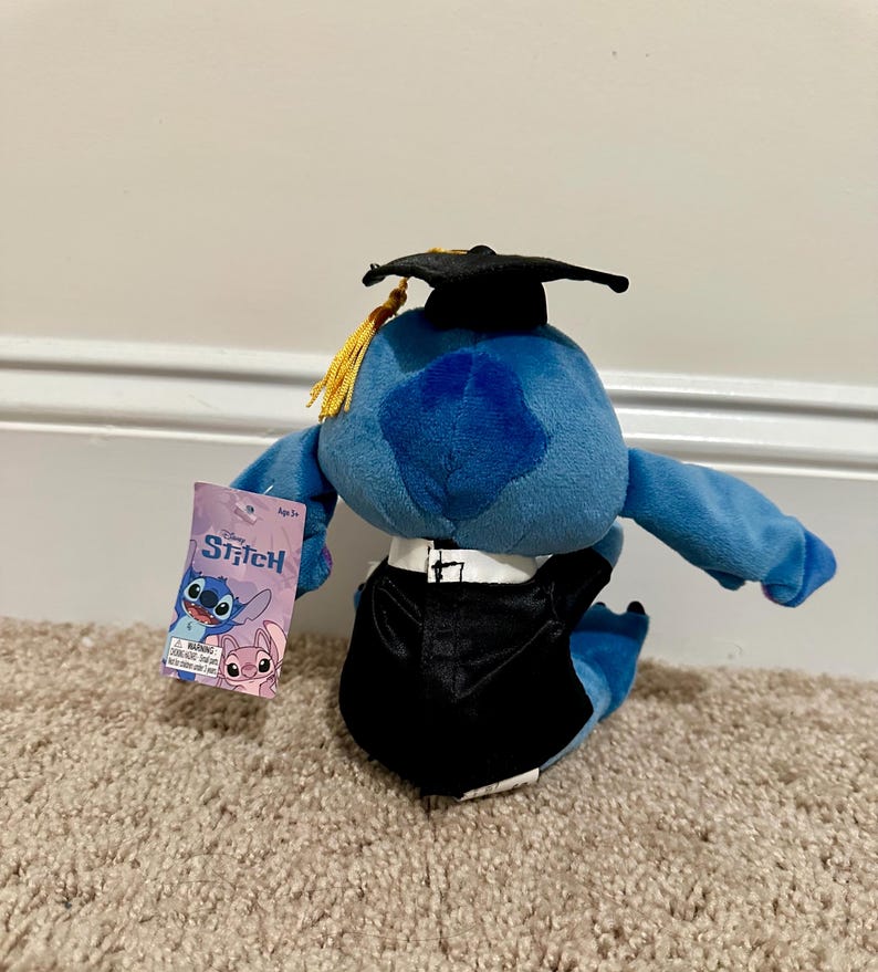 May include: A blue Stitch plush toy dressed in a black graduation gown and mortarboard with a gold tassel. The toy holds a tag with the Stitch logo and images of Stitch and Angel.