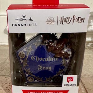 Może przedstawiać: Ozdoba choinkowa Hallmark w pudełku. Pudełko jest czerwono-białe, z napisami "Chocolate Frog Box" i "Harry Potter". Ozdoba ma kształt pięciokąta z napisem "Chocolate Frog".