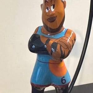 May include: A ceramic ornament depicting a cartoon character in a blue and orange basketball uniform. The figure has crossed arms, a brown complexion, and red shoes. The number 6 is visible on the uniform.