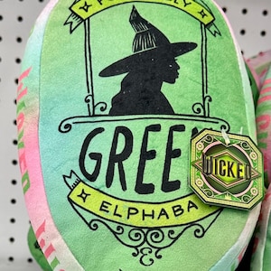 Peut inclure: Coussin en peluche vert et rose avec le texte "Positively Green Elphaba Wicked" et une silhouette de sorcière portant un chapeau. Le coussin porte une étiquette indiquant "Wicked".