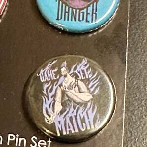 K&ouml;nnte beinhalten: Ein Set von Ansteckbuttons mit Cartoon-Illustrationen und Text. Ein Button ist t&uuml;rkis mit dem Wort "DANGER" in Wei&szlig;. Ein weiterer Button ist schwarz mit den Worten "GAME SET MATCH" und einer Charakterillustration.