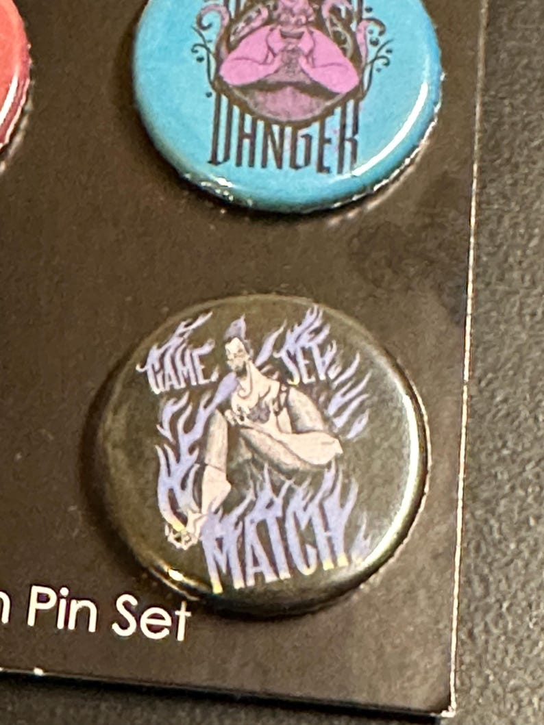 K&ouml;nnte beinhalten: Ein Set von Ansteckbuttons mit Cartoon-Illustrationen und Text. Ein Button ist t&uuml;rkis mit dem Wort "DANGER" in Wei&szlig;. Ein weiterer Button ist schwarz mit den Worten "GAME SET MATCH" und einer Charakterillustration.