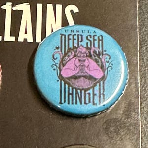 K&ouml;nnte beinhalten: Ein runder, hellblauer Knopf mit dem Disney Villains-Logo und dem Text "URSULA DEEP SEA DANGER". Er zeigt eine lila Illustration von Ursula, der Meerhexe, aus Arielle, die Meerjungfrau.
