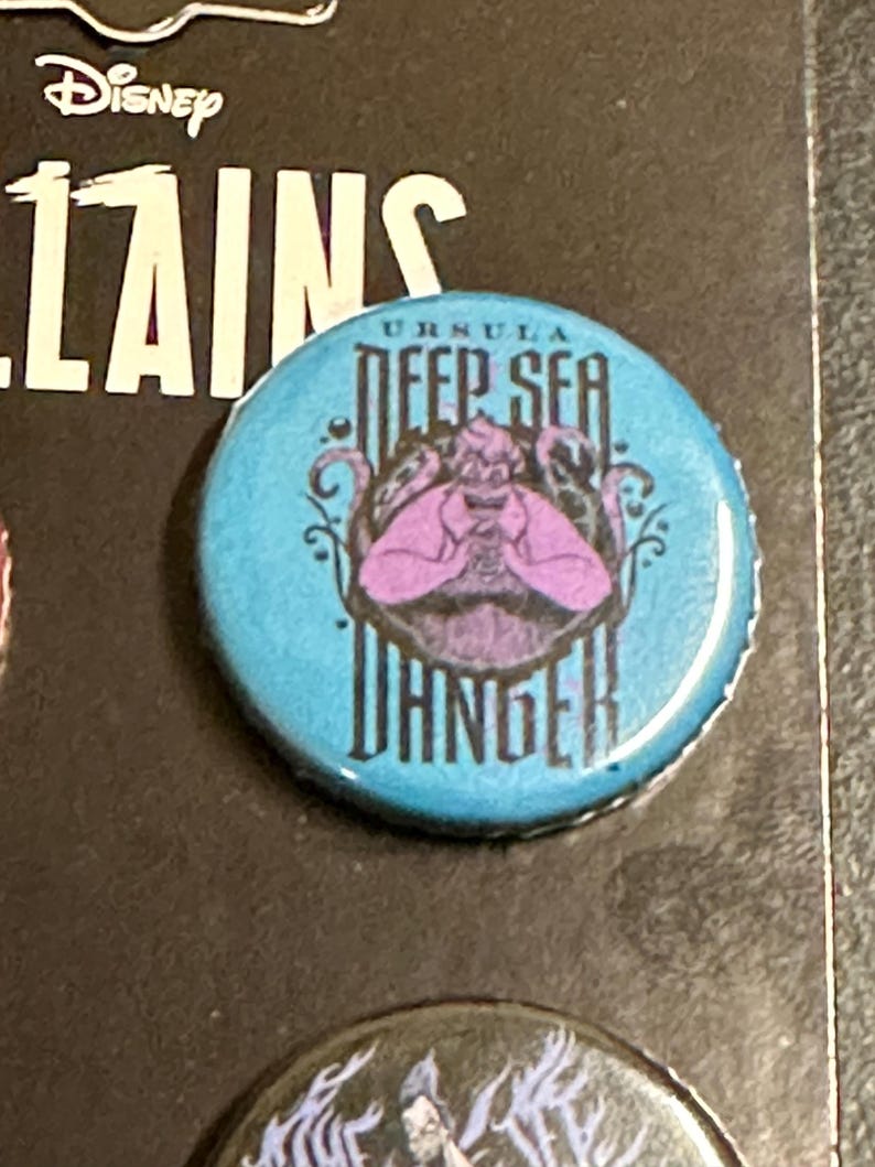 K&ouml;nnte beinhalten: Ein runder, hellblauer Knopf mit dem Disney Villains-Logo und dem Text "URSULA DEEP SEA DANGER". Er zeigt eine lila Illustration von Ursula, der Meerhexe, aus Arielle, die Meerjungfrau.