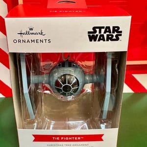 May include: A Star Wars TIE Fighter Christmas tree ornament in a box. The gray and blue fighter is in a clear plastic case. The box is white with the Hallmark and Star Wars logos. Text on the box reads "TIE FIGHTER" and "CHRISTMAS TREE ORNAMENT".