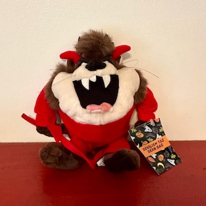 May include: A plush toy of the Tasmanian Devil, dressed in a red devil costume with horns. The toy has a white face, large teeth, and a black open mouth. A tag reads "Devilish Taz Bean Bag".