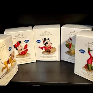 Op de afbeelding: Een verzameling Hallmark Keepsake Disney-ornamenten in hun originele dozen. De dozen bevatten personages als Mickey Mouse, Minnie Mouse, Donald Duck en Goofy. Elk ornament is een miniatuurbeeldje, perfect voor de feestdagen.