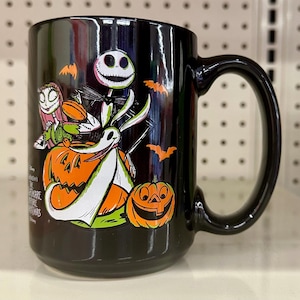 May include: A black ceramic mug featuring a colorful graphic of Jack Skellington, Sally, and Zero from The Nightmare Before Christmas. The design includes orange pumpkins, small orange bats, and the movie title text printed in white on the side.