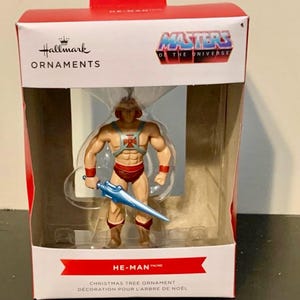 Può includere: Un ornamento per albero di Natale Hallmark in scatola con He-Man di Masters of the Universe. La figura è muscolosa, indossa slip rossi, stivali e polsini, con una spada blu. La scatola recita "HE-MAN" e "ORNAMENTO PER L'ALBERO DI NATALE".