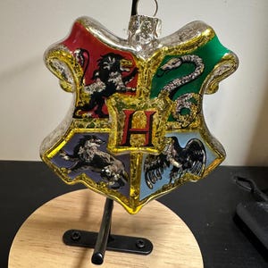 May include: A glass Christmas ornament in the shape of a shield, featuring the Hogwarts crest. The crest is divided into four sections with a lion, snake, badger, and eagle. The ornament has red, green, blue, and gold accents.