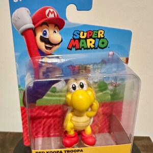 Può includere: Una action figure di Super Mario di un Koopa Troopa giallo in una confezione di plastica trasparente. La figura ha scarpe rosse e un guscio. La confezione presenta Mario e le parole "Super Mario".
