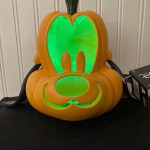 May include: An orange plastic pumpkin-shaped light featuring the carved face of Pluto with long black ears. The interior glows with a bright green light, illuminating the cutouts for the eyes and nose. A small green stem sits on top of the pumpkin.