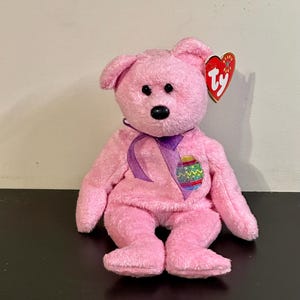 May include: A pink plush bear toy with a purple ribbon and an Easter egg design. The bear has black eyes and a red heart-shaped tag. A classic children's toy.