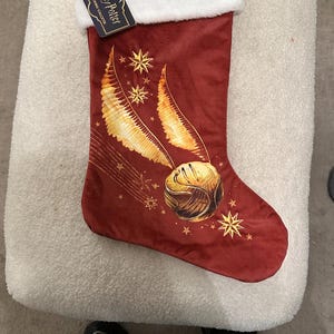 Peut inclure: Une chaussette de Noël bordeaux avec un revers blanc, brodée de "Harry Potter" en or. La chaussette présente un design de vif d'or et d'étoiles dorées. Une étiquette avec le logo Harry Potter est attachée.