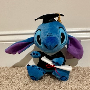 May include: A blue Stitch soft toy, dressed in a graduation cap and gown, clutching a rolled diploma tied with a red ribbon. The toy has large black eyes, purple-lined ears, and sits on a carpet.