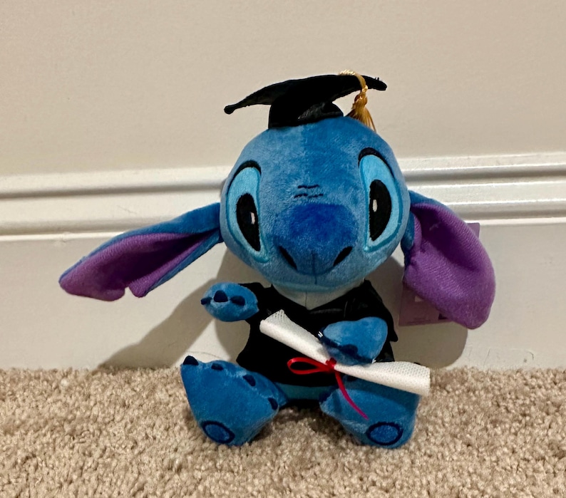 May include: A blue Stitch soft toy, dressed in a graduation cap and gown, clutching a rolled diploma tied with a red ribbon. The toy has large black eyes, purple-lined ears, and sits on a carpet.