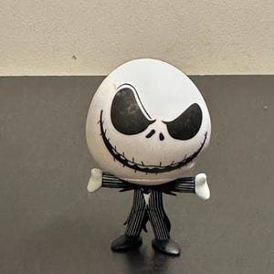 May include: A figurine of Jack Skellington from The Nightmare Before Christmas. The figure has a white head with black eyes and a stitched grin. He wears a black and white pinstripe suit with a bat-shaped bow tie and black shoes.