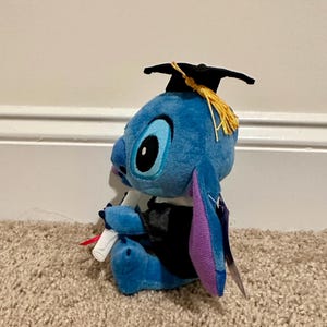 May include: A blue plush Stitch doll wearing a black graduation cap with a gold tassel and a black graduation gown. The doll is holding a rolled-up diploma with a red ribbon. The plush toy has large black eyes and purple inner ears.