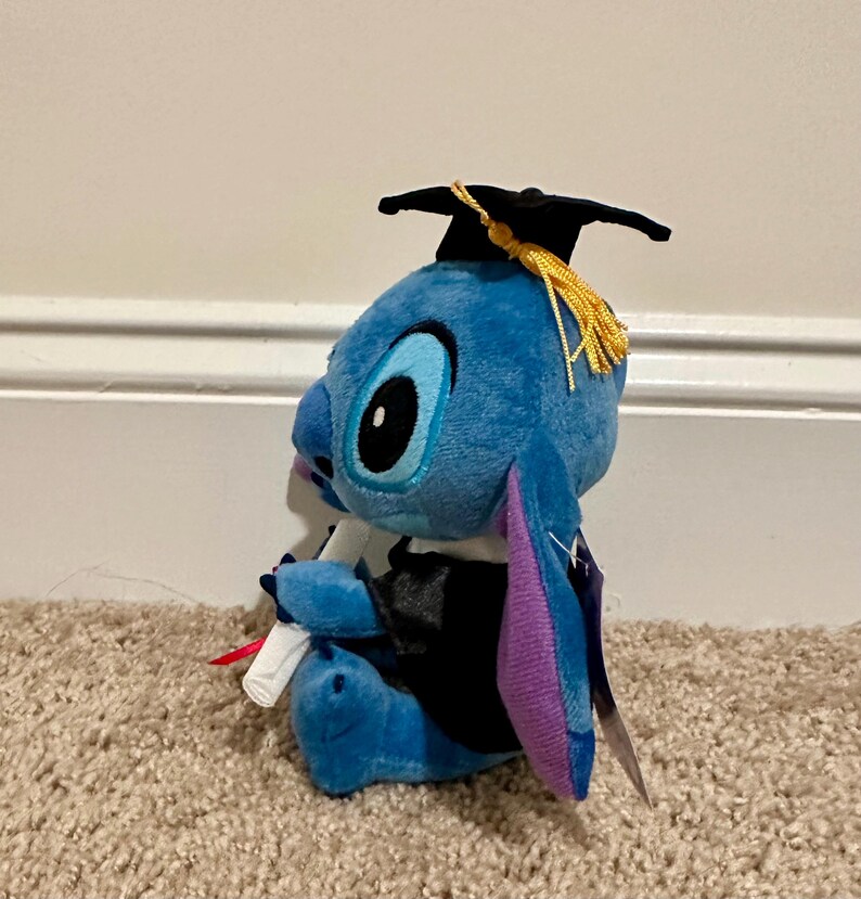 May include: A blue plush Stitch doll wearing a black graduation cap with a gold tassel and a black graduation gown. The doll is holding a rolled-up diploma with a red ribbon. The plush toy has large black eyes and purple inner ears.