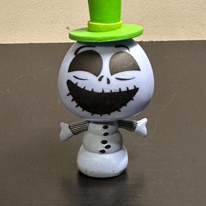 May include: A light blue figurine of a snowman character with a smiling face and a green top hat. The snowman has black eyes and a stitched mouth. The arms are striped black and white. The snowman has three buttons down the front.
