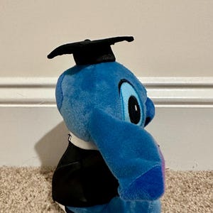 May include: A blue plush toy of a cartoon character wearing a black graduation cap and gown. The toy has large eyes and ears, and is sitting on a beige carpet. The character is a popular Disney character.
