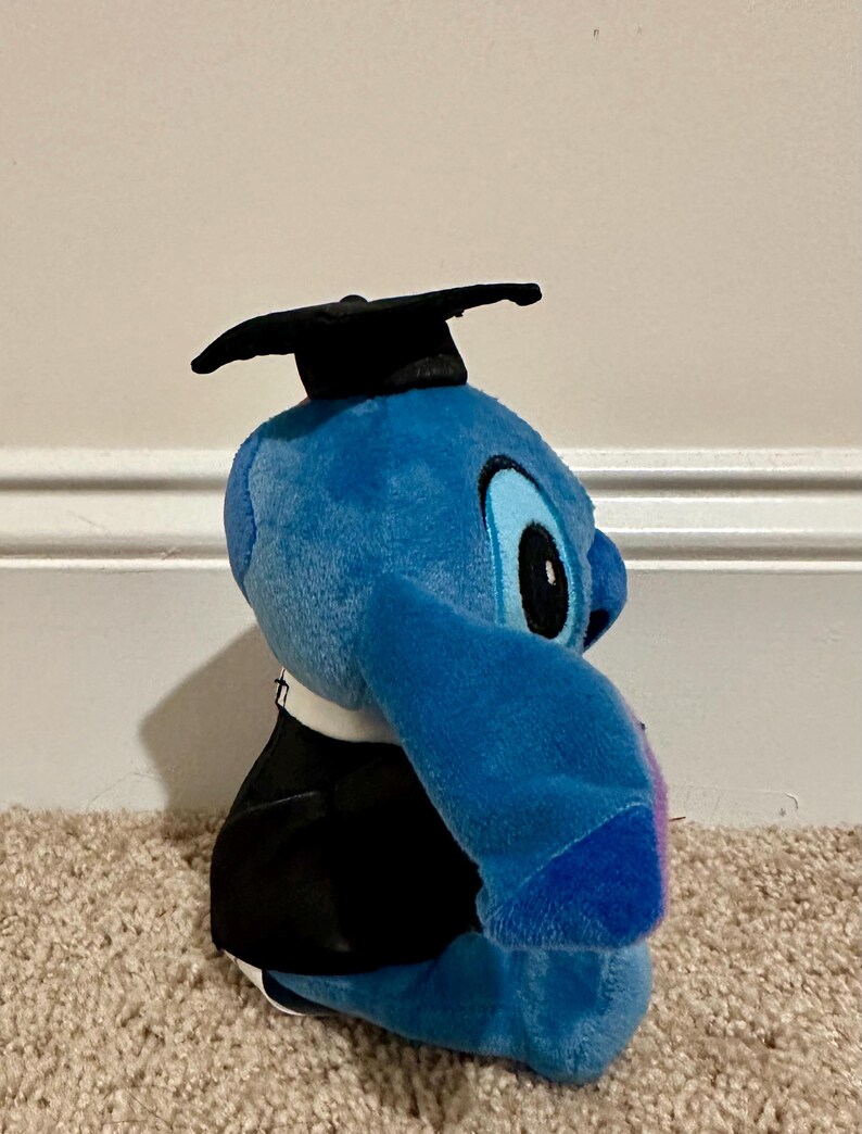 May include: A blue plush toy of a cartoon character wearing a black graduation cap and gown. The toy has large eyes and ears, and is sitting on a beige carpet. The character is a popular Disney character.