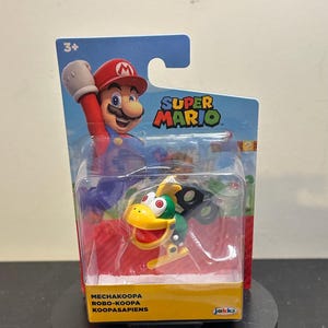 Może przedstawiać: Figurka Super Mario Mechakoopa Robo-Koopa Koopasapiens, zapakowana w przezroczyste plastikowe etui. Opakowanie przedstawia Mario i napisy "Super Mario". Figurka jest zielona, żółta i czarna.