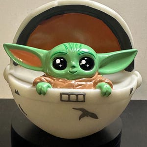 May include: A green figurine of a character from a popular science fiction series, with large eyes and pointed ears, seated in a white pod-like carrier. The carrier has black accents and a bird-like symbol.