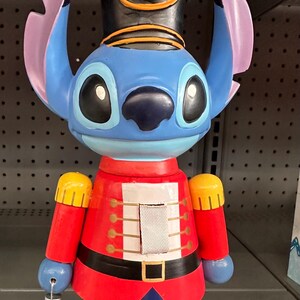 Puede incluir: Figura de cascanueces de Stitch azul. La figura tiene una cara azul, orejas grandes y lleva una chaqueta roja con detalles dorados, un sombrero negro y una bola verde. La figura está sobre una base roja.