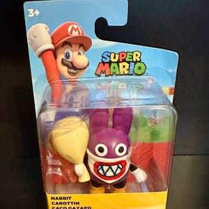 May include: A Super Mario action figure set in a clear plastic package. The set includes a Mario figure with a red hat and a purple rabbit-like character with a wide grin. The packaging features the "Super Mario" logo and the text "Nabbit, Carottin, Caco Gazapo, Mopsie, Ruboniglio."