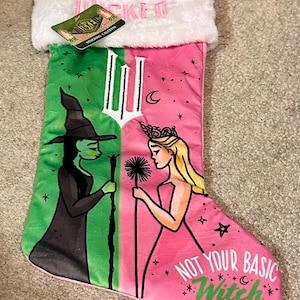 Puede incluir: Un calcetín navideño con un diseño verde y rosa que presenta ilustraciones de dos personajes. Una es una bruja de piel verde y la otra es una princesa rubia. El texto "NOT YOUR BASIC Witch" está en el calcetín.