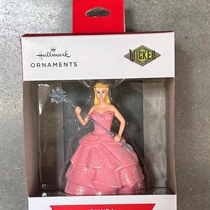May include: A Hallmark Christmas tree ornament featuring Glinda from Wicked. The figurine wears a pink gown and holds a wand. The box reads "Hallmark Ornaments" and "Christmas Tree Ornament".
