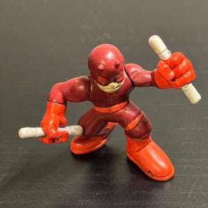 May include: A red and maroon Daredevil action figure with white batons. The figure is wearing a red mask, gloves, and boots. The figure is in a fighting stance, holding a baton in each hand.