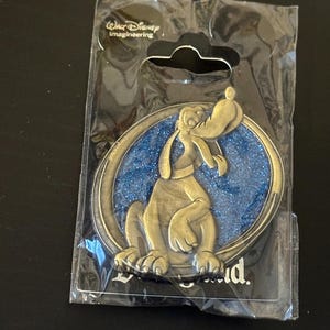 May include: A gold-colored pin featuring the Disney character Goofy, set against a blue glitter background within a circular frame. The pin is in its original packaging, with the "Walt Disney Imagineering" logo visible.
