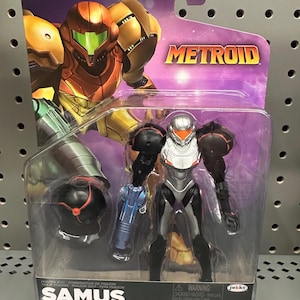 Op de afbeelding: Een Samus-actiefiguur in een Phazon-pak, verpakt in een doorzichtige plastic blisterverpakking. Het figuur heeft een zilveren en zwart harnas met een oranje vizier en een blauw armkanon. De verpakking bevat een Morph Ball-accessoire en Metroid-branding.