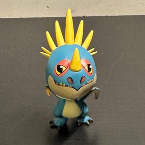 May include: A blue and yellow dragon figurine with a spiky head crest and a yellow horn. The dragon has large eyes with red accents and a friendly expression. The figurine is a collectible toy.