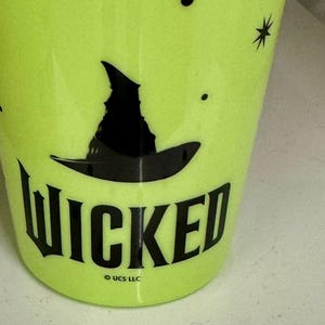 May include: A neon green tumbler with the word "WICKED" in black, along with a black witch hat graphic. The tumbler also features small black star and dot designs. The bottom of the tumbler has the text "© UCS LLC".