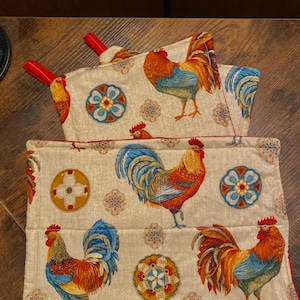 May include: Set of three beige kitchen pot holders with red trim. The pot holders feature a rooster design with colorful accents and a repeating pattern of circular designs.