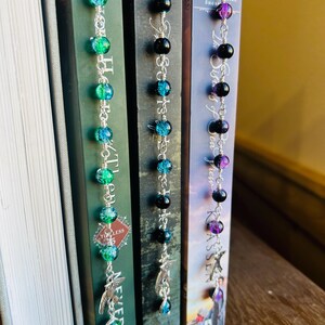 May include: Three beaded bookmarks with silver chains and charms are displayed next to books. The bookmarks feature colorful beads in shades of green, blue, black, and purple. Each bookmark has a unique charm, including a bird and a teardrop.