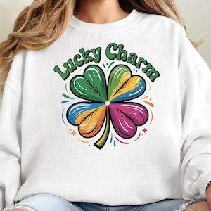 Pode incluir: Camisola branca com um design colorido de trevo de quatro folhas. O trevo é segmentado em seções verdes, amarelas, azuis e rosas. As palavras "Lucky Charm" estão arqueadas acima do trevo em verde.