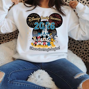 May include: White sweatshirt featuring a colorful graphic design. The design includes the text "Disney Family Vacation 2026" with images of Mickey Mouse, Minnie Mouse, Donald Duck, Goofy, and a castle. The phrase "Where the magic begins!" is also included.