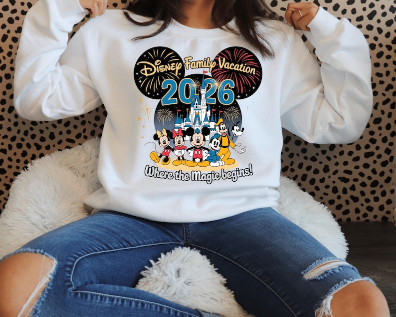 May include: White sweatshirt featuring a colorful graphic design. The design includes the text "Disney Family Vacation 2026" with images of Mickey Mouse, Minnie Mouse, Donald Duck, Goofy, and a castle. The phrase "Where the magic begins!" is also included.