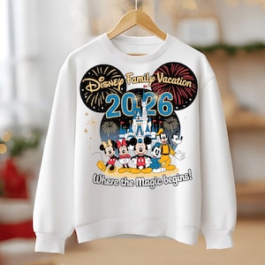 May include: White sweatshirt featuring a Disney Family Vacation design for 2026. The graphic includes Mickey, Minnie, Donald, Goofy, and a castle, with the text "Where the Magic begins!" and fireworks.
