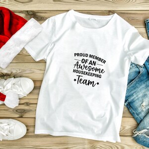 May include: White t-shirt with black text that reads "Proud Member of an Awesome Housekeeping Team". The shirt is laid out on a wooden surface with a red Santa hat and blue jeans.