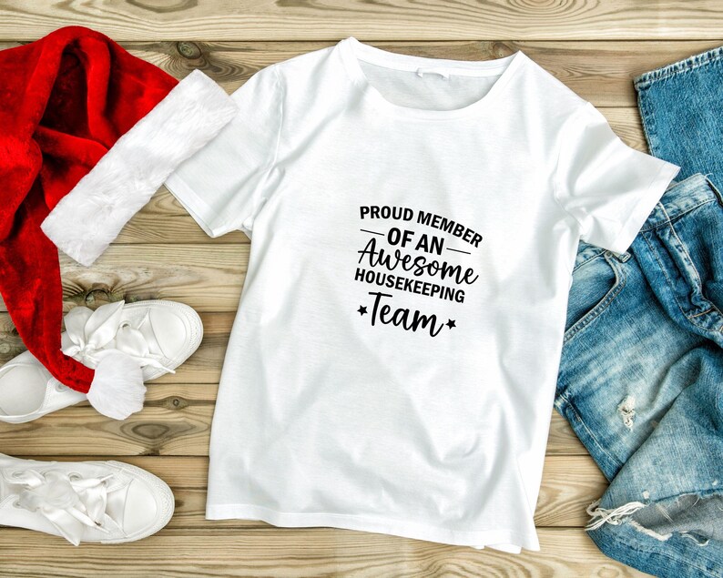 May include: White t-shirt with black text that reads "Proud Member of an Awesome Housekeeping Team". The shirt is laid out on a wooden surface with a red Santa hat and blue jeans.