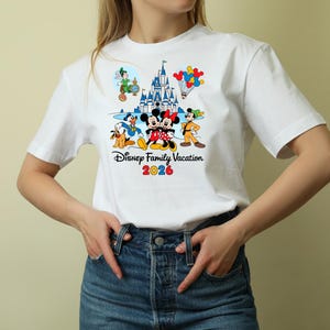 May include: White t-shirt featuring a colorful graphic of Disney characters, including Mickey and Minnie Mouse, Donald Duck, and Peter Pan, with a castle and balloons. The text "Disney Family Vacation 2026" is printed below the characters.