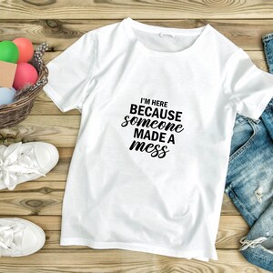 May include: White t-shirt with black text that reads "I'm here because someone made a mess". The shirt is laid out on a wooden surface with a basket of colourful Easter eggs and a pair of blue jeans.
